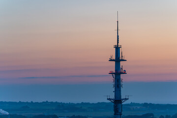 Obraz premium A tall transmission tower rises against a pastel evening sky. The horizon stretches in soft orange and pink hues.