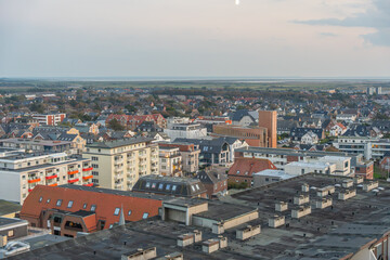 Aerial view over a coastal town with houses, apartments, and fields stretching to the horizon. The evening light casts a soft glow.