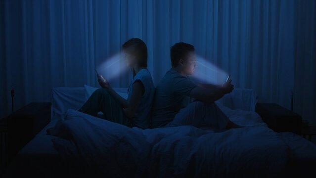 A couple sits back to back in bed at night using smartphones
