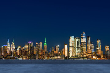 Rooftop view of illuminated Manhattan skyline at night with modern skyscrapers and iconic Empire State Building under dark blue sky in New York City, USA.