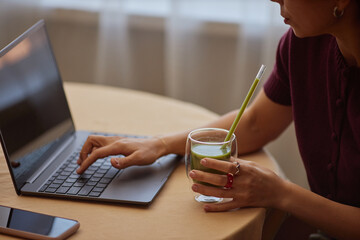 Young adult Caucasian woman working on laptop while holding glass of green smoothie with straw, sitting at round table, focusing on screen, hand typing on keyboard