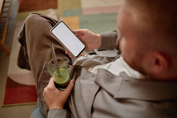 Caucasian young adult man sitting and holding smartphone with blank screen in one hand while drinking green smoothie with reusable straw, relaxing in living room with patterned rug