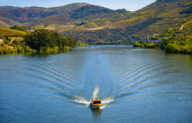 View from the old steel truss bridge “Ponte do Pinhão” over the river in the idyllic Douro Valley in northern Portugal. A small motorboat approaches the bridge. Wide angle scenery and waterway.