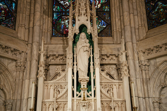 This image features a detailed white marble statue of the Virgin Mary holding the Christ Child, set within a richly carved altar niche at St. Patrick's Cathedral in New York City. Stained glass