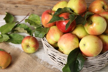 Fresh ripe apples with green leaves in wicker basket on white wooden table, closeup