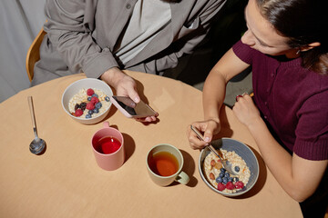 Young adult Caucasian man using smartphone, while sitting with young adult Caucasian woman eating oatmeal with berries at table with cups of tea and coffee during breakfast