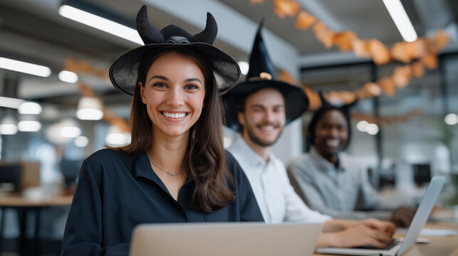 A cheerful team of coworkers wearing playful Halloween accessories such as witch hats devil horns and skeleton masks while working on laptops in a bright open plan office