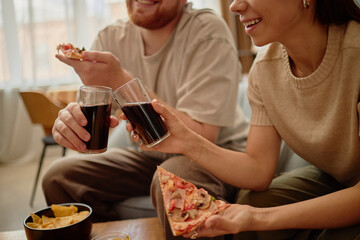 Caucasian young man and Caucasian young woman sitting together eating pizza and drinking soda, smiling and interacting, holding food and beverages in relaxed indoor setting
