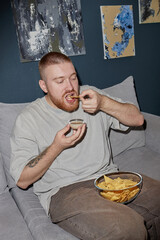 Caucasian young adult man sitting on sofa eating tortilla chips and dipping chip into sauce, holding small bowl in one hand, casual setting with abstract paintings on wall