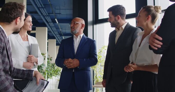 Leader talking to business team on meeting in modern glass office. Senior businessman explaining ideas to corporate colleagues, standing via open discussion, people listening speaker on briefing