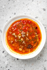 Homemade beef and vegetable soup in a ceramic bowl on light countertop. Rich tomato broth with potatoes, carrots, herbs. Rustic comfort food, top view, natural lighting