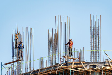 Three construction workers are working on the top of a building under construction against a clear blue sky. They are surrounded by steel rebar, and one worker is shown tying the rebar together.