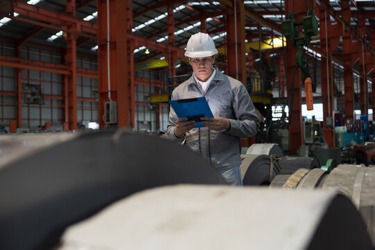 Metalwork manufacturing, warehouse of raw materials. Male factory worker inspecting quality rolls of metal sheet in factory during manufacturing process, wearing safety uniform - Powered by Adobe