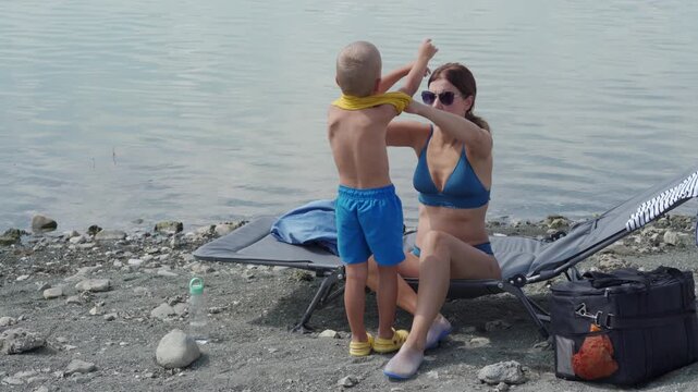 A young boy removes his yellow shirt while his mother, in a blue bikini, sits on a beach chair beside him on a rocky lakeside shore