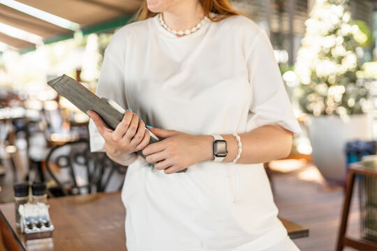 A young Caucasian woman with long brown hair holds a tablet in a cafe. She wears a white shirt and a smartwatch. The setting is bright and modern, ideal for remote work. - Powered by Adobe