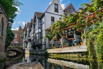 Ivy draped over the gazebo roof of a riverside restaurant and a wattle and daub rendered building with multicolour stained glass square windows all seen from the river Stour in Canterbury, Kent UK. 
