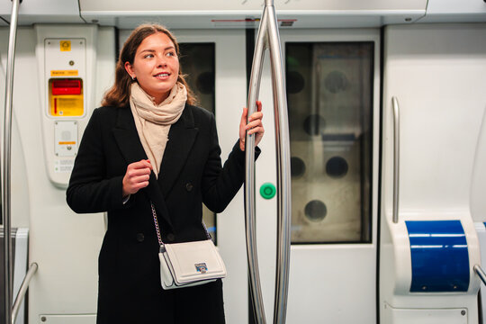 A young woman stands inside a subway train, holding onto a pole with a thoughtful expression. She gazes around the moving vehicle, engaged in her commute, showcasing the daily life of a city passenger