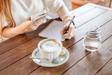 A young woman with long red hair writes in a notebook while holding a smartphone. A cup of cappuccino and a glass of water are on the wooden table.