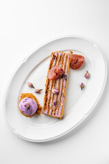 Gourmet plated dessert with layered pastry, purple cream, fig, flower garnish, and berry ice cream on crumb base. Elegant presentation on white dish, fine dining concept.