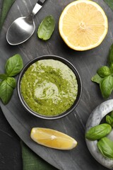 Tasty green pesto in bowl, basil leaves and lemon on black table, top view