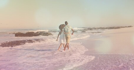 Strolling African American couple arm-in-arm into surf at sandy beach, with waves and dark rocks