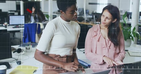 Two office colleagues working together at shared desk in open plan office, with laptop and tablet