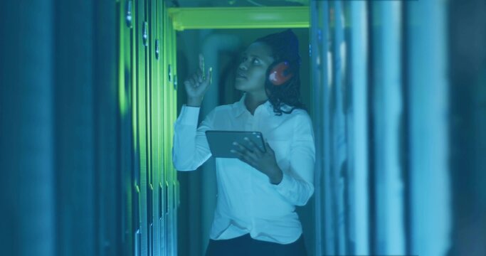 Inspecting female IT technician adjusting server module in data center aisle with tablet, earmuffs