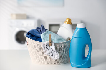 Laundry basket with baby clothes and bottles of detergent on white table indoors