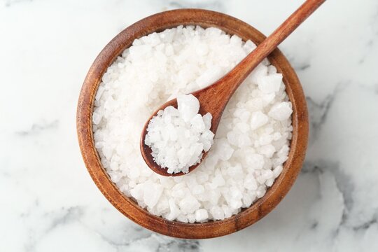 Sea salt in bowl and spoon on white marble table, top view