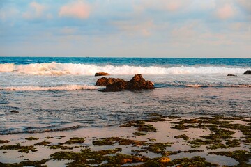 Horizontal landscape of white ocean waves breaking over a dark rock formation on an algae-covered tidal flat, under a pastel sunset sky.