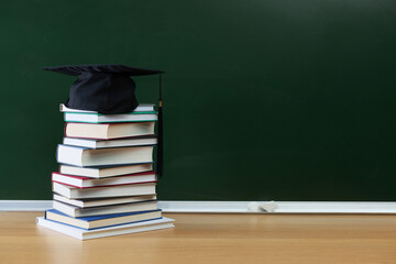 Happy Teacher's Day. Stack of books and mortarboard on wooden table near chalkboard, space for text