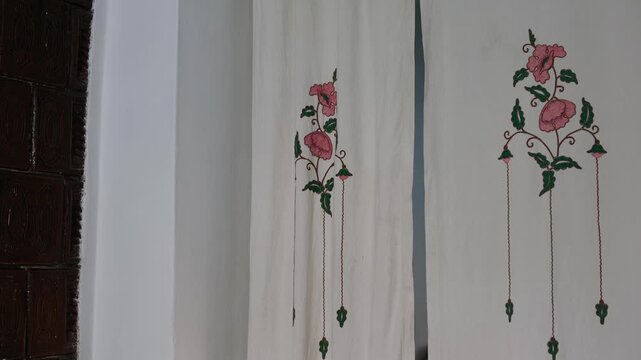 Hand embroidered curtain with pink floral pattern hangs between doors in old rural house, soft light and simple rustic interior create cozy village atmosphere.
