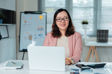 Obraz premium Confident Asian businesswoman working at her desk with a laptop, smiling towards the camera. Business and career concept