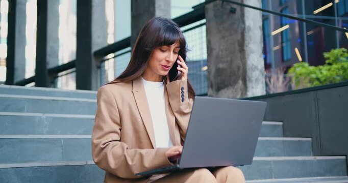 Smiling businesswoman sitting on outdoor stairs, working on laptop and talking on smartphone. Confident female professional working remotely in urban setting