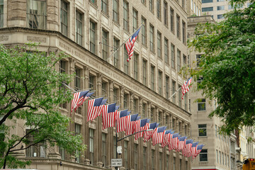 Multiple American flags are displayed in rows along the facade of a historic New York City building. The scene highlights patriotism and classic architecture surrounded by green trees in a downtown