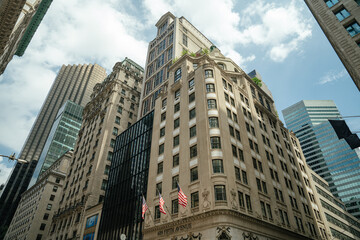 A striking upward view of Fifth Avenue shows historic buildings with American flags alongside sleek glass skyscrapers. The contrast of old and new architecture under a bright blue sky reflects the