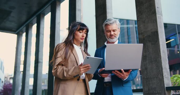 Two business partners, a man and a woman in formal attire, are standing outside a modern office building, discussing work-related topics using a digital tablet and a laptop