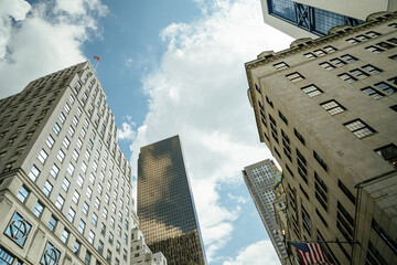 A view looking up at a group of Manhattan skyscrapers blending modern glass towers with historic stone buildings. The American flag waves at the top of the frame beneath a bright blue sky with