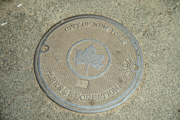 A close-up view of a manhole cover from New York City Parks and Recreation shows a maple leaf emblem. The textured metal surface is surrounded by concrete pavement in a public urban area.