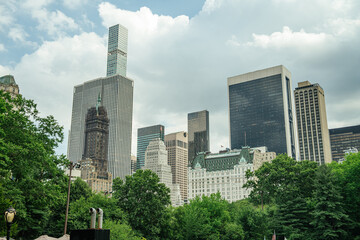 A mix of classic and modern skyscrapers stands tall behind dense green trees in Midtown Manhattan. The iconic skyline includes historic hotels and sleek towers under a cloudy sky.