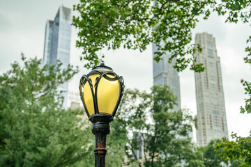 A classic yellow street lamp stands among green trees in Central Park, New York City. Tall modern skyscrapers blur in the background, creating a mix of old and new urban elements.