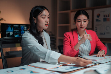 Fototapeta premium Two young Asian businesswomen working late in the office, discussing financial data and analyzing charts during a meeting