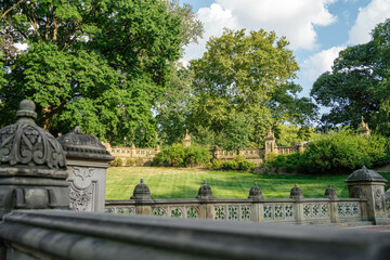 Ornate stone railings frame a grassy area surrounded by lush trees in Central Park, New York. The historic architecture contrasts beautifully with the vibrant summer landscape.