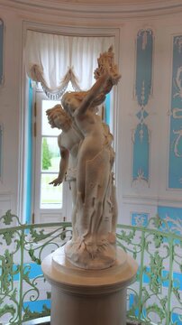 A majestic vertical full shot captures a white marble classical sculpture of two intertwined mythological figures positioned atop a pedestal on a grand interior staircase landing. The statue is