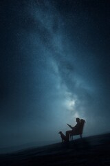 A man is sitting in an armchair in the middle of a mountainside and reading next to his dog under the starry night sky with the Milky Way against the background of a peaceful remote landscape