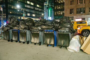 A row of trash bins overloaded with black garbage bags lines the sidewalk in Manhattan. The background reveals busy city streets, lit storefronts, and a yellow taxi passing by at night.