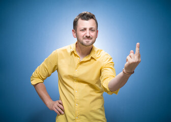Smiling man giving the middle finger - showing fuck off gesture over blue background, dresses in yellow shirt. One guy with obscene gesture and looking to camera