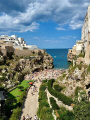 Sommerlicher Strand in Polignano a Mare, Apulien &ndash; malerische Felsk&uuml;ste mit t&uuml;rkisblauem Meer, wei&szlig;en H&auml;usern und vielen Badeg&auml;sten unter dramatischem Himmel.