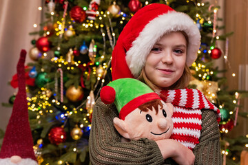 Excited Child in Santa Hat with Christmas Tree Background holding a festive gnome toy, smiling brightly in front of a beautifully lit and decorated Christmas tree. magical holiday ambiance