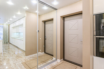 A modern hallway with beige walls, tiled floor, a full-length mirror, and a door. A glimpse of a white kitchen can be seen
